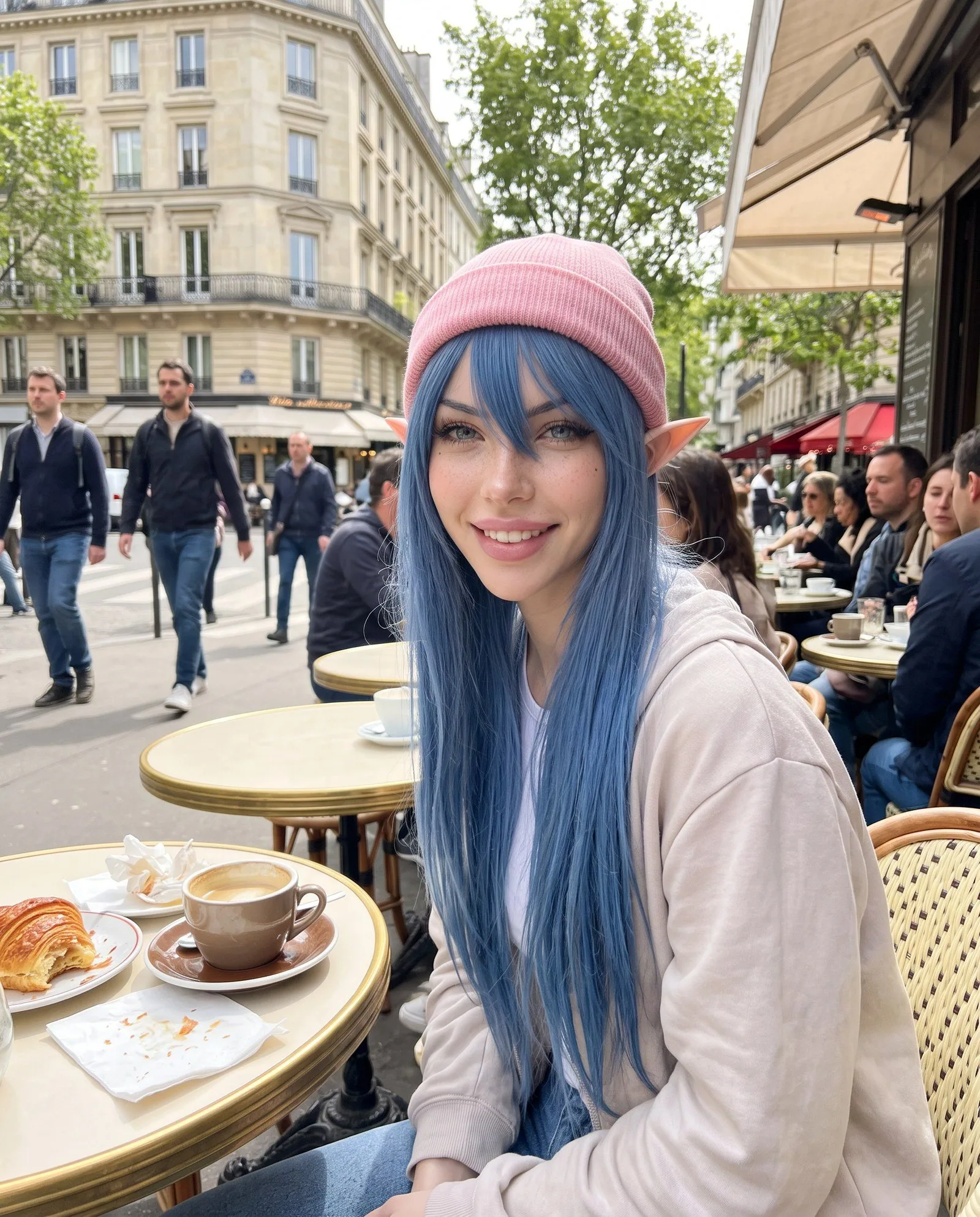Photo verticale façon téléphone, en plein jour : White Rose, longs cheveux bleus raides avec frange, bonnet rose, oreilles pointues d'elfe, sweat à capuche beige, sourit à l'objectif, assise à une terrasse de café parisienne ; sur la petite table ronde, un café et un croissant ; derrière elle, des passants, des clients attablés et un immeuble haussmannien.