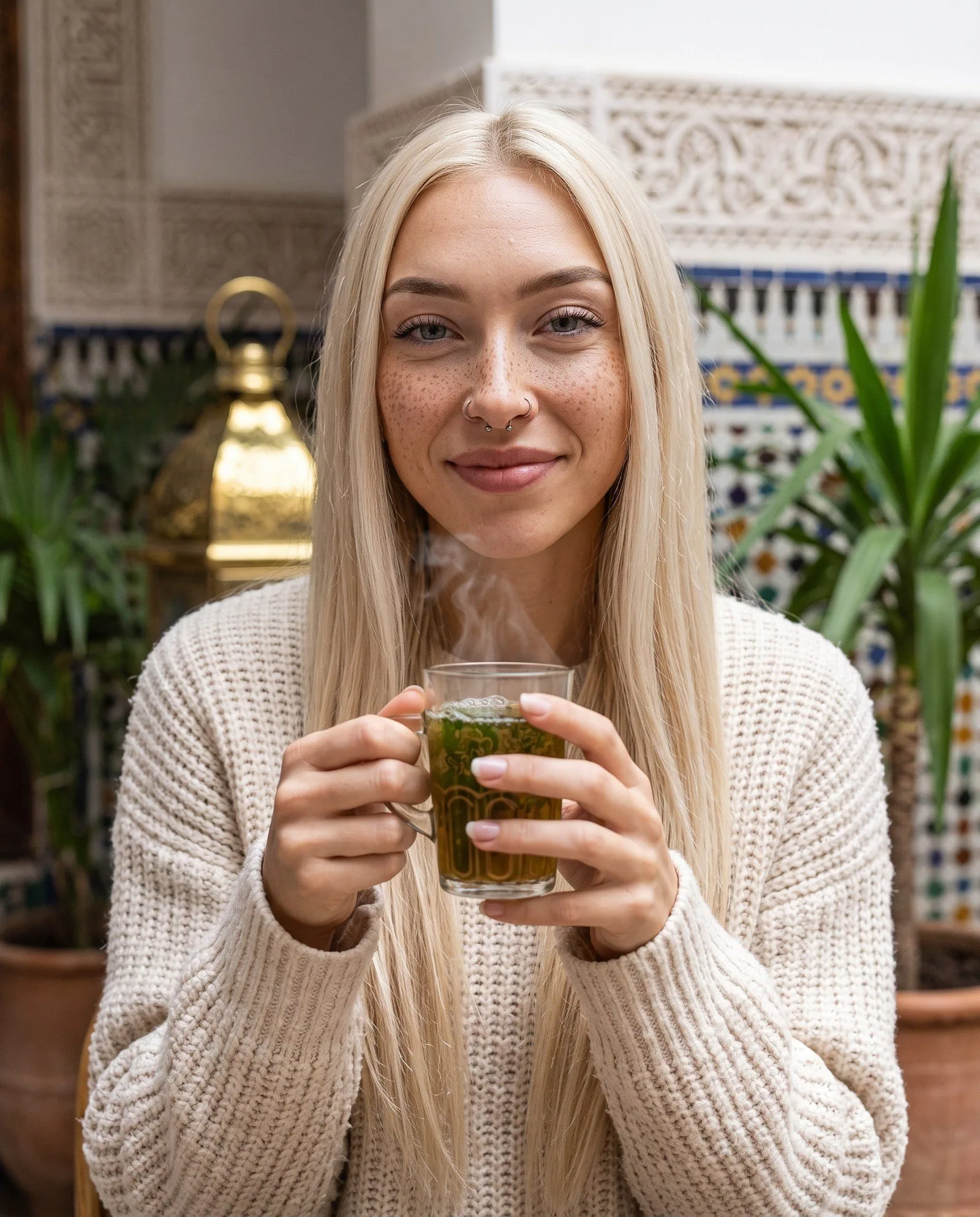 Portrait vertical : Kira, longs cheveux blond platine, taches de rousseur, un anneau à chaque narine et un au septum, pull en grosse maille crème, tient à deux mains un verre de thé à la menthe fumant et sourit légèrement ; derrière elle, plâtre sculpté, carreaux de zellige, une lanterne dorée et des plantes en pot.