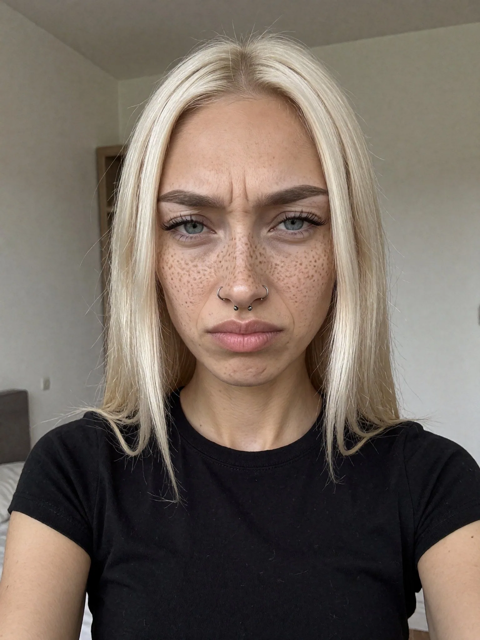 Selfie vertical en intérieur : Kira, cheveux blond platine, taches de rousseur, un anneau à chaque narine et un au septum, t-shirt noir, fronce fort les sourcils et fixe l'objectif ; derrière elle, des murs blancs et un coin de lit à gauche.