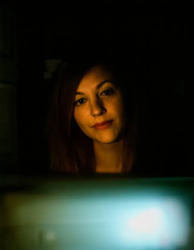 Femme dans une cuisine sombre, visage éclairé par en dessous, grande tache lumineuse floue au premier plan.