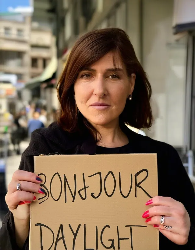 Femme en manteau noir, dans une rue, qui tient un carton où on lit « BONJOUR », B mal formé et J à l'envers, puis « DAYLIGHT ».